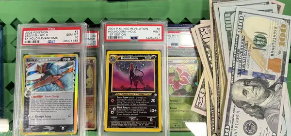 A vendor case at a card show: rows of PSA graded Pokemon cards standing on green grid shelving, a gem mint Espeon and a gem mint Scizor from Undaunted on the top row, a gem mint Deoxys from Holon Phantoms and a mint first edition Houndoom from Neo Revelation below, and a folded stack of hundreds and fifties resting against the glass beside them.